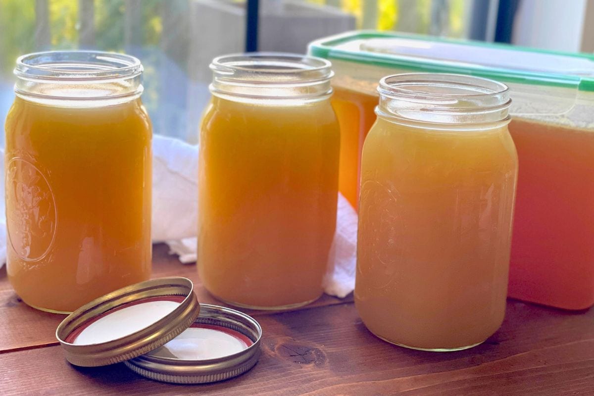 Several jars of homemade broth in varying shades of amber to golden hues, lined up on a wooden surface with lids placed beside them.