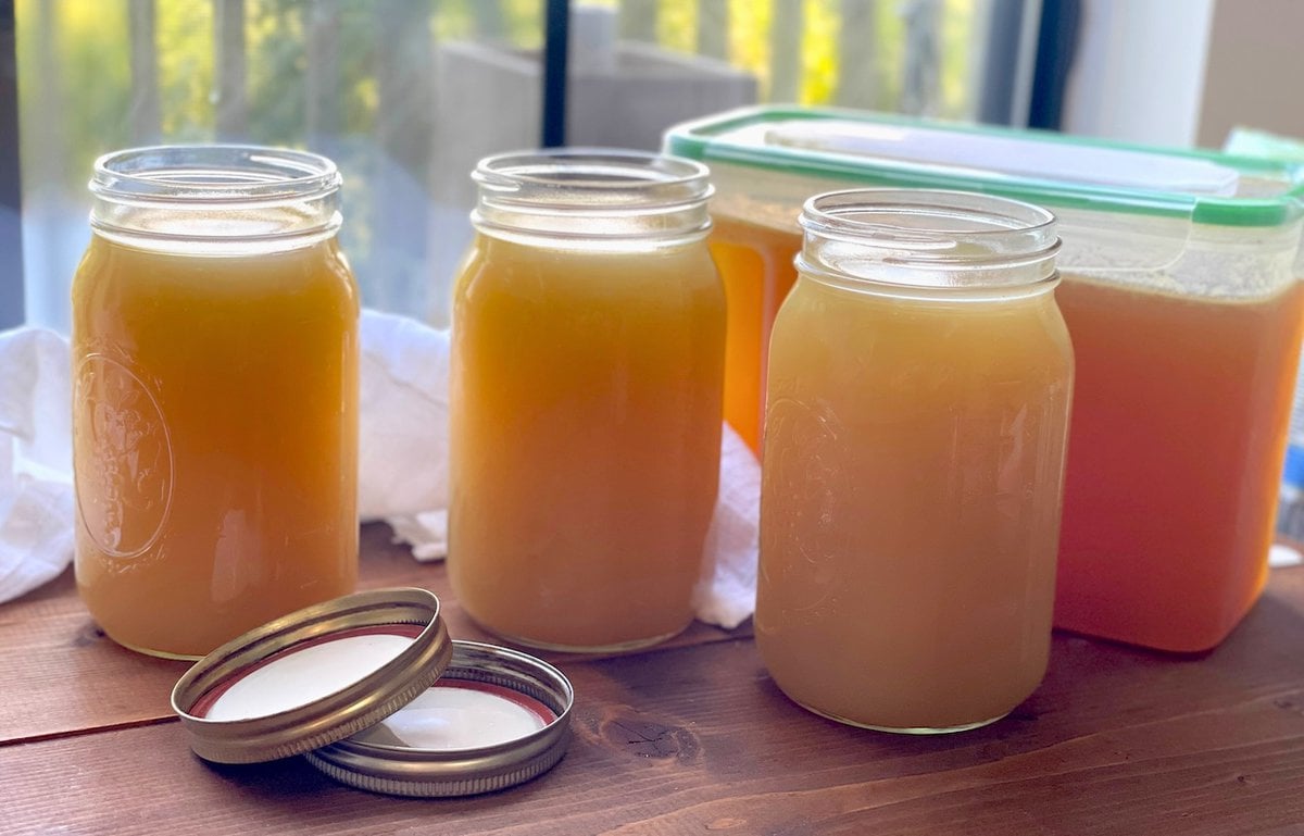 Four jars of pressure cooker turkey broth on a wooden surface with lids placed beside them, against a backdrop of a balcony view.