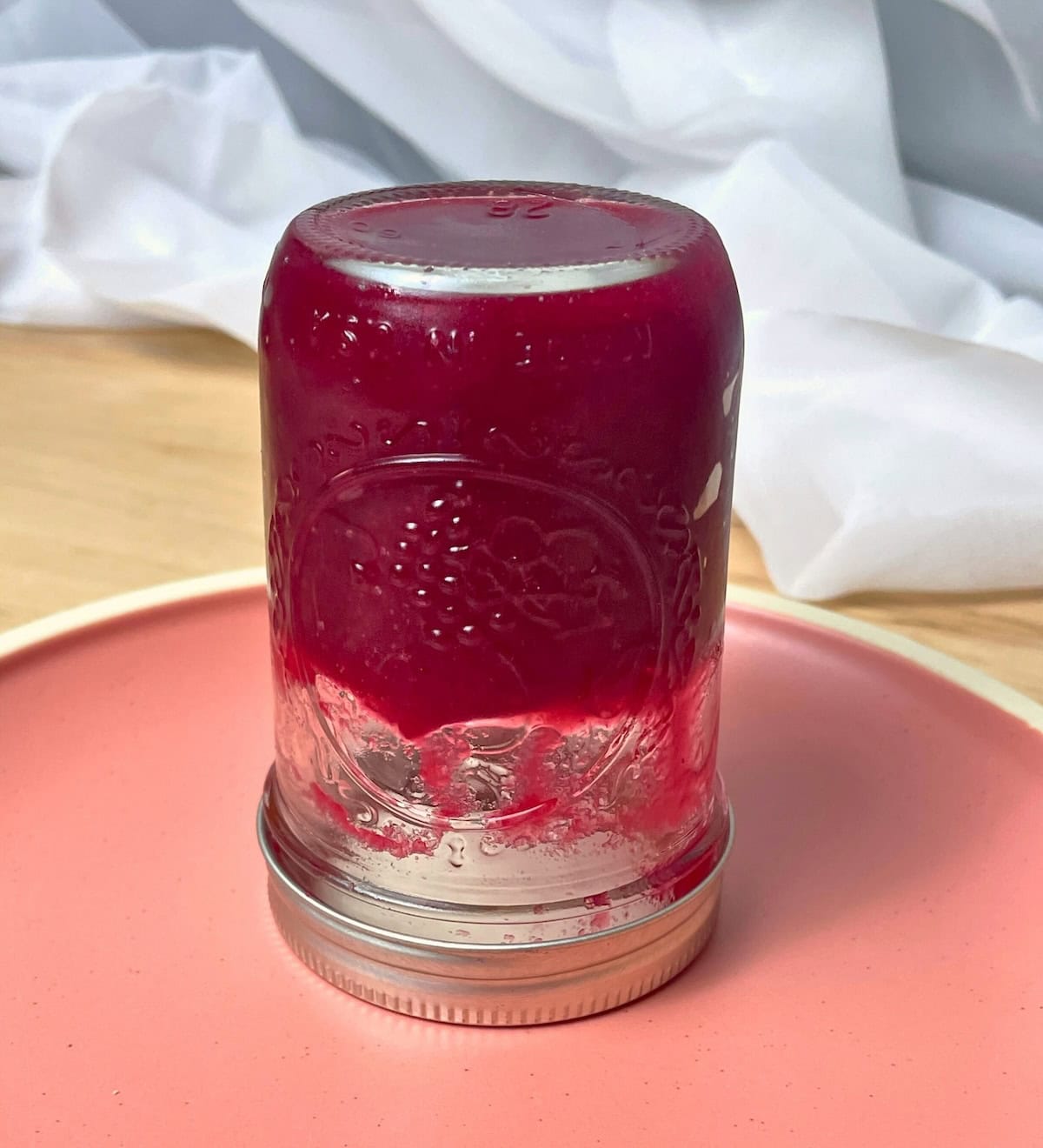 A glass jar of cranberry sauce turned upside down on a pink plate, with a crumpled white cloth in the background.