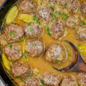 Close-up view of savory meatballs topped with grated cheese and herbs in gravy, served with pickled peppers, in a cast iron skillet with a wooden spoon.