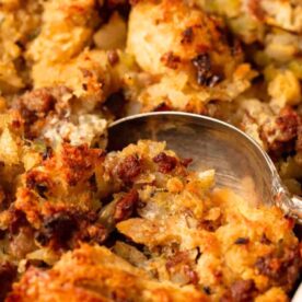 A close-up view of a baked bread stuffing in a white casserole dish with a spoon, showing crispy edges and moist interior.