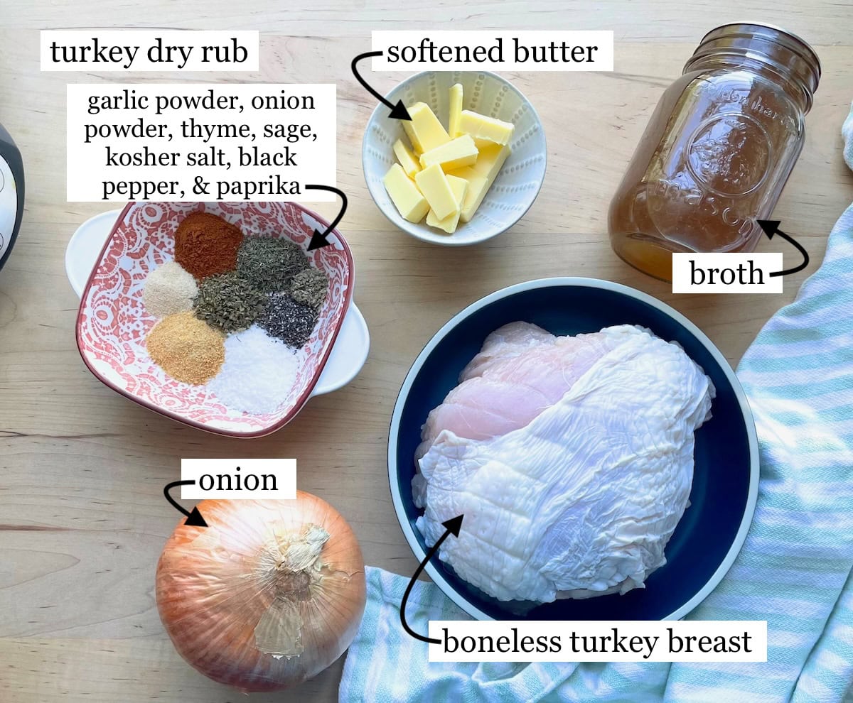 A countertop view of ingredients for seasoning a turkey breast, including a bowl of mixed spices, softened butter, broth, and a raw boneless turkey breast.