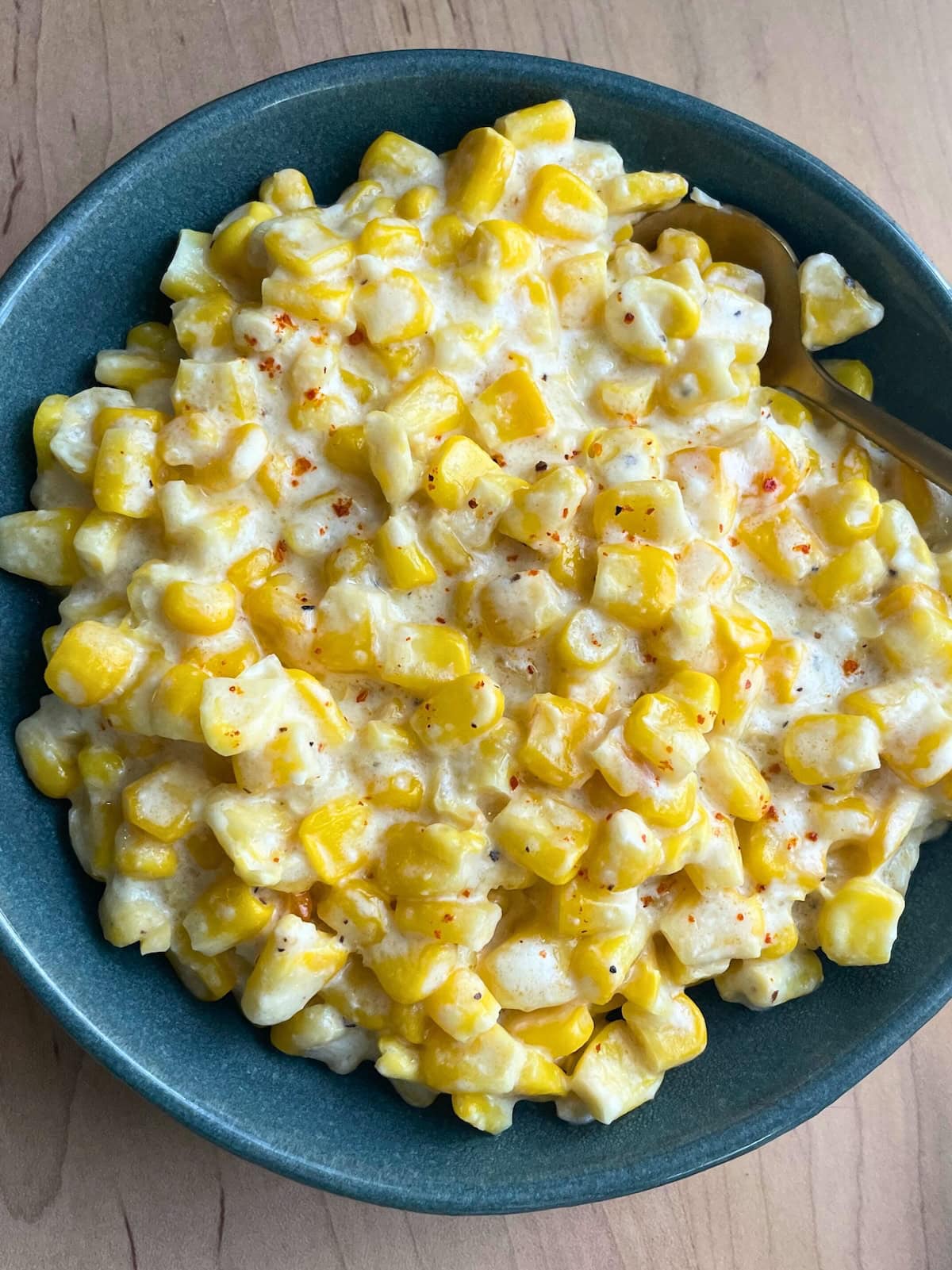 A bowl of creamed corn with a sprinkle of red pepper on a wooden table, next to a floral cloth napkin.