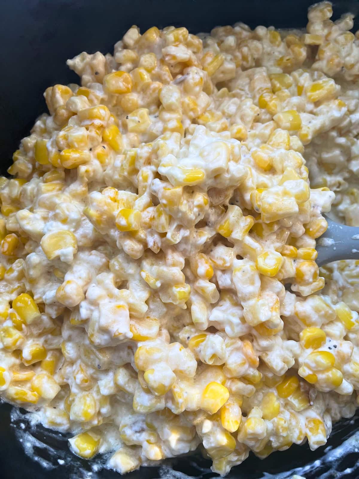 A detailed view of a creamy corn casserole in a black crockpot with a spoon, showcasing the texture and ingredients.