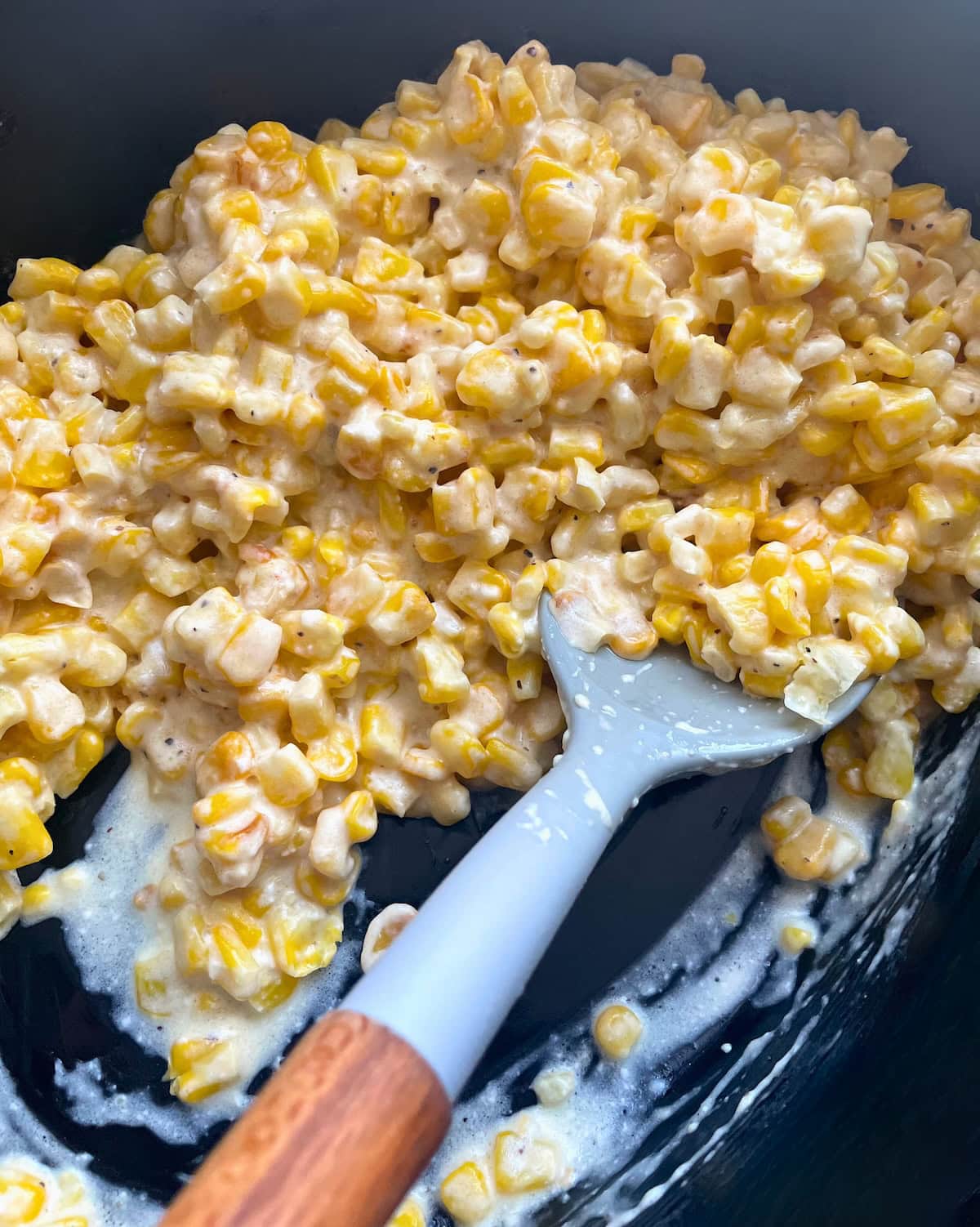 Close-up view of creamy corn being stirred with a gray spatula in a black crock pot.