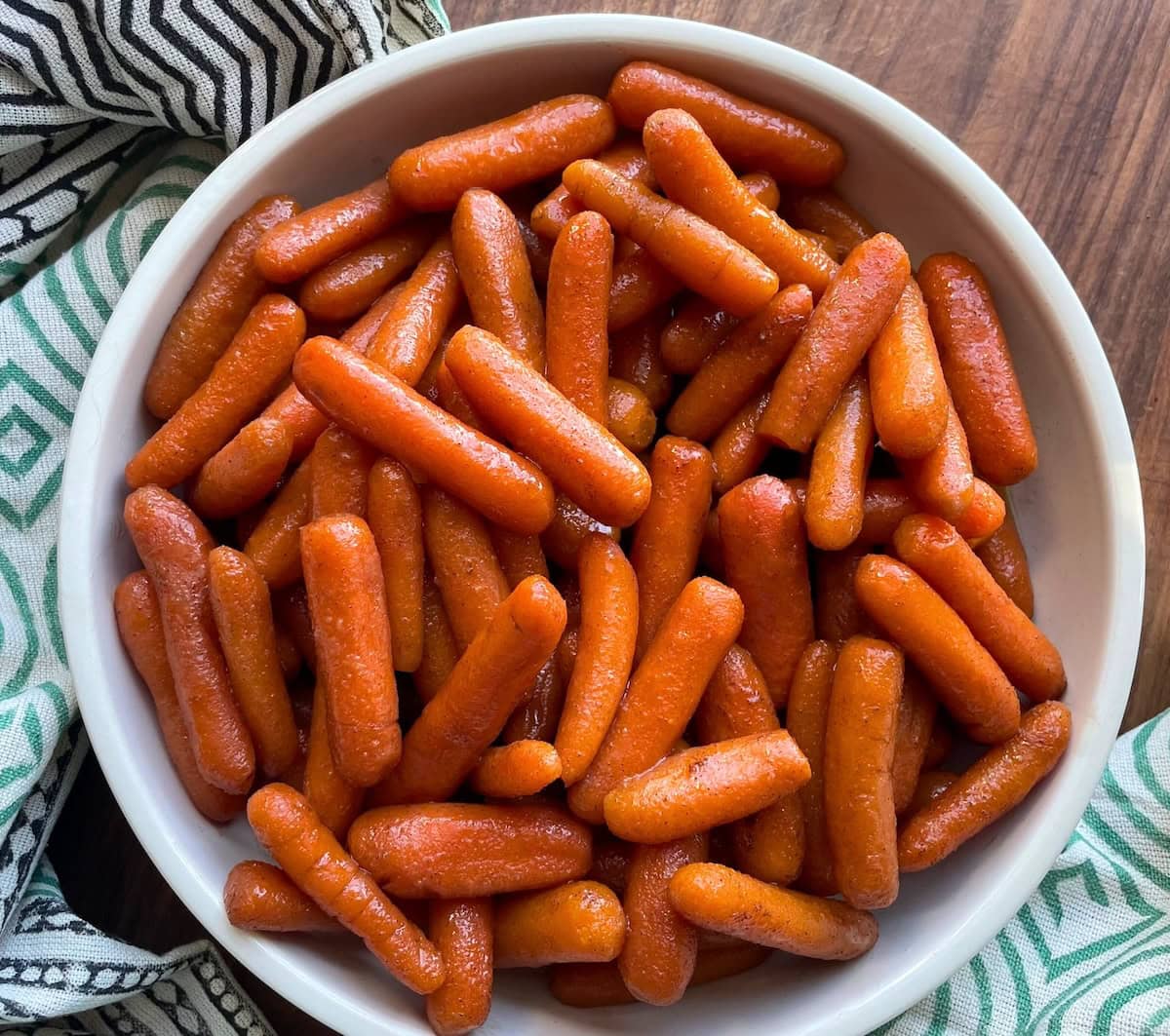 A white bowl filled with shiny glazed baby carrots on a wooden table, with a patterned cloth napkin alongside.