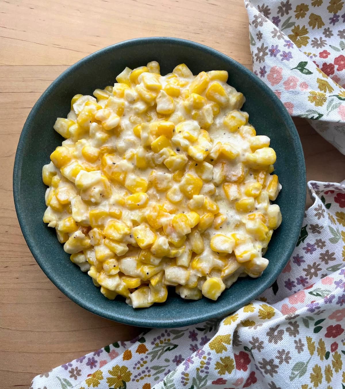 A bowl of creamed corn with a sprinkle of red seasoning on a wooden table, next to a floral cloth napkin.