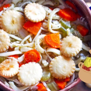 A close-up of homemade chicken noodle soup in a blue-handled pot with crackers on top, garnished with freshly ground black pepper.