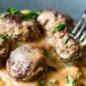 Close-up of meatballs with gravy on mashed potatoes, garnished with chopped parsley and a fork piercing one meatball.