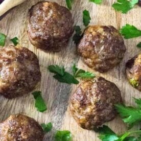 Succulent homemade meatballs garnished with parsley leaves on a rustic wooden cutting board, accompanied by a purple cloth napkin to the side.