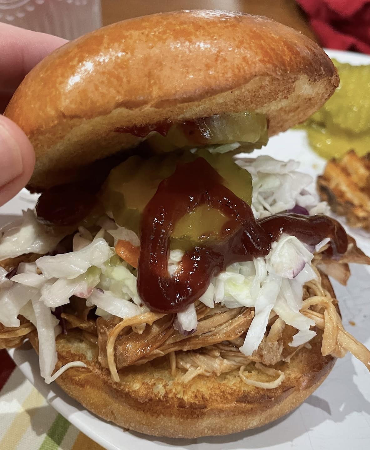 Close-up view of a hand holding a pulled chicken sandwich with coleslaw, pickles, and sauce.