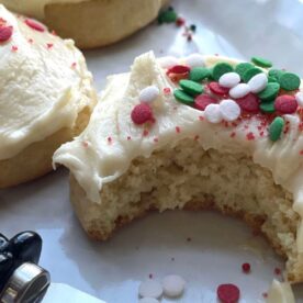 A close-up of sugar cookies topped with white frosting and colorful holiday sprinkles, with one cookie partially bitten into, revealing a soft interior.