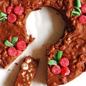 A chocolate-covered cake shaped like a wreath, garnished with red and green candied fruit, partially sliced.