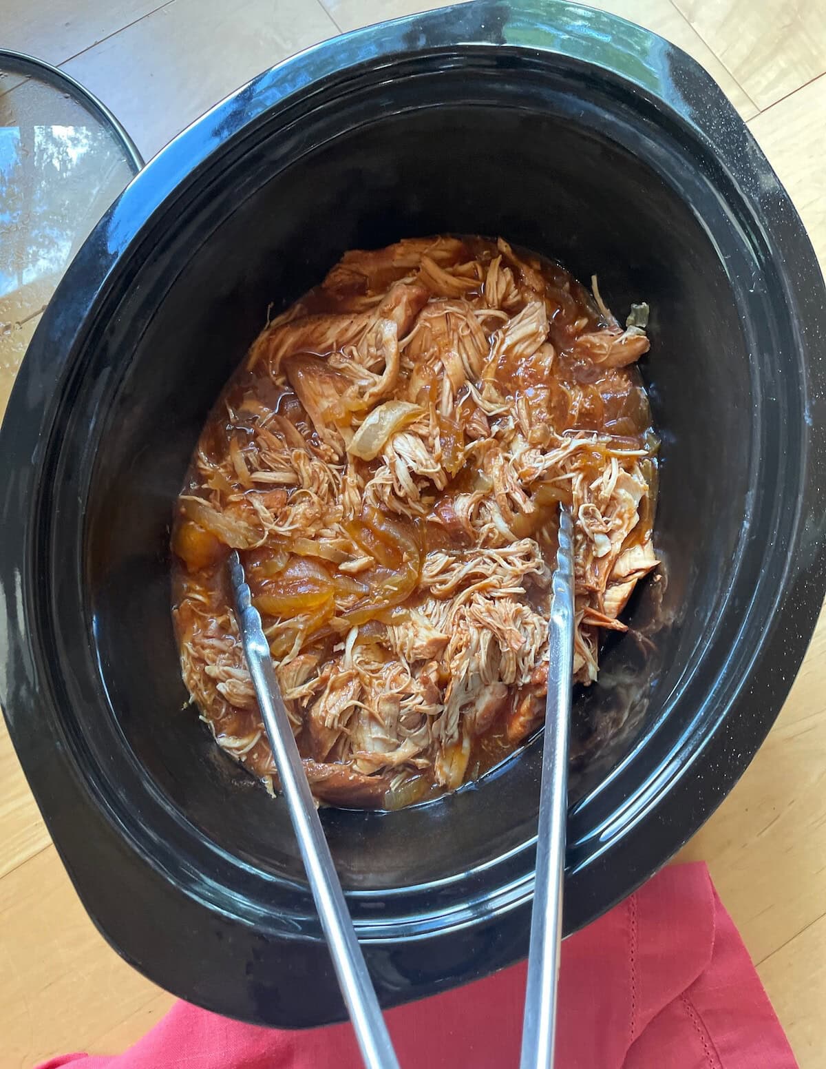 A top-down view of shredded pulled chicken in a black crock pot with tongs, resting on a wooden surface with a red cloth underneath.