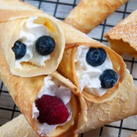 Close-up of homemade krumkakes with cream and topped with blueberries and a raspberry, dusted with powdered sugar on a cooling rack.