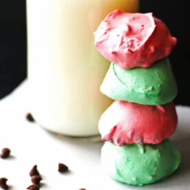 A stack of pastel-colored cookies in pink, green, and blue next to a glass of milk, with chocolate chips scattered around on a white surface.