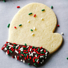 A mitten-shaped sugar cookie partially dipped in chocolate and decorated with colorful sprinkles on a light grey background.