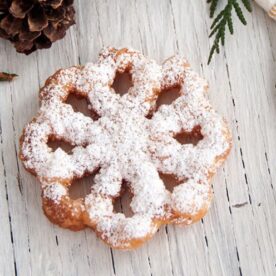 A snowflake-shaped rosette covered with powdered sugar on a rustic wooden background, accompanied by green foliage and a pine cone.