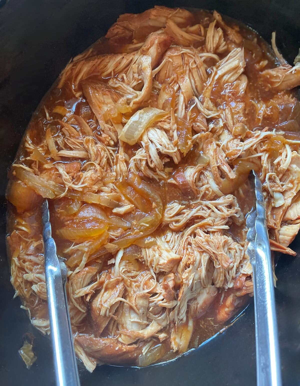 A close-up view of shredded barbecue chicken in a rich sauce, served in a black crockpot with metal tongs.