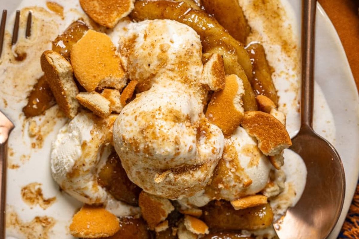 A close-up of a dessert featuring baked apples topped with vanilla ice cream and crushed cookies, drizzled with caramel sauce.