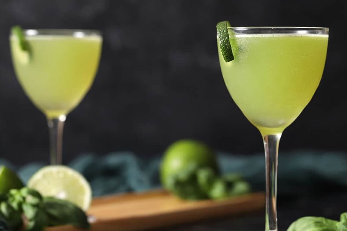 A close-up of two vibrant green cocktails garnished with lime slices, surrounded by fresh limes and herbs on a wooden surface against a dark background.