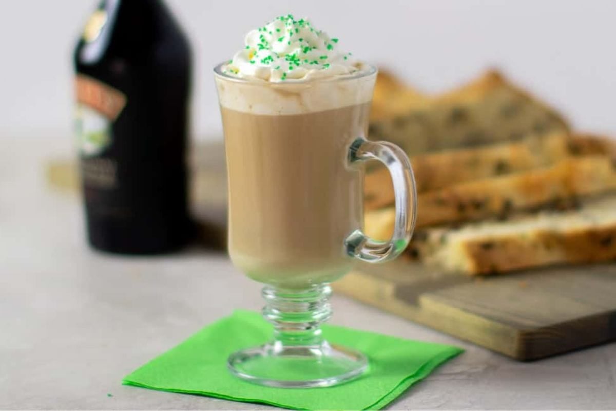 A glass of creamy Irish coffee topped with whipped cream and festive green sprinkles, set on a green napkin, with baked treats blurred in the background.