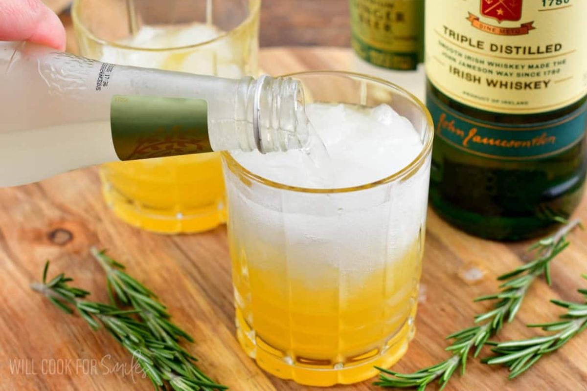 A hand pouring ginger beer into a glass of ice and whiskey, garnished with fresh rosemary, set on a wooden surface.