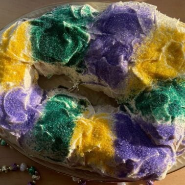 A colorful, decorated king cake featuring green, purple, and yellow sugar, displayed on a glass plate next to a beaded crown and necklace.