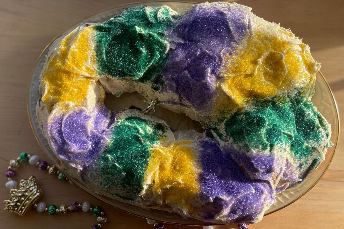 A colorful, decorated king cake featuring green, purple, and yellow sugar, displayed on a glass plate next to a beaded crown and necklace.