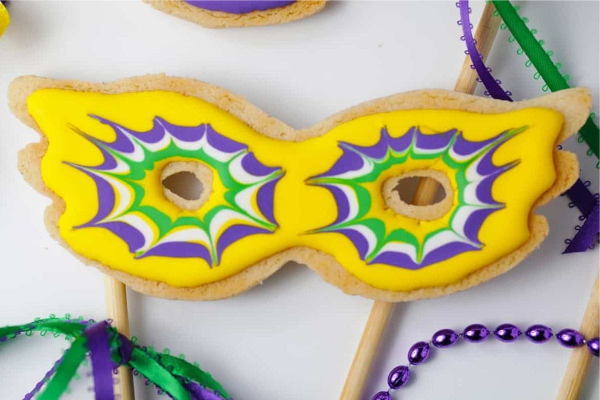 A vibrant yellow masquerade mask-shaped cookie decorated with purple, green, and white icing, surrounded by festive ribbons and beads.