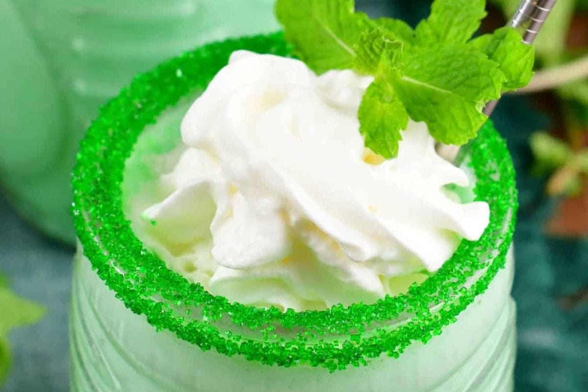 Close-up of a minty green beverage topped with whipped cream and garnished with mint leaves, featuring a green sugar rim.