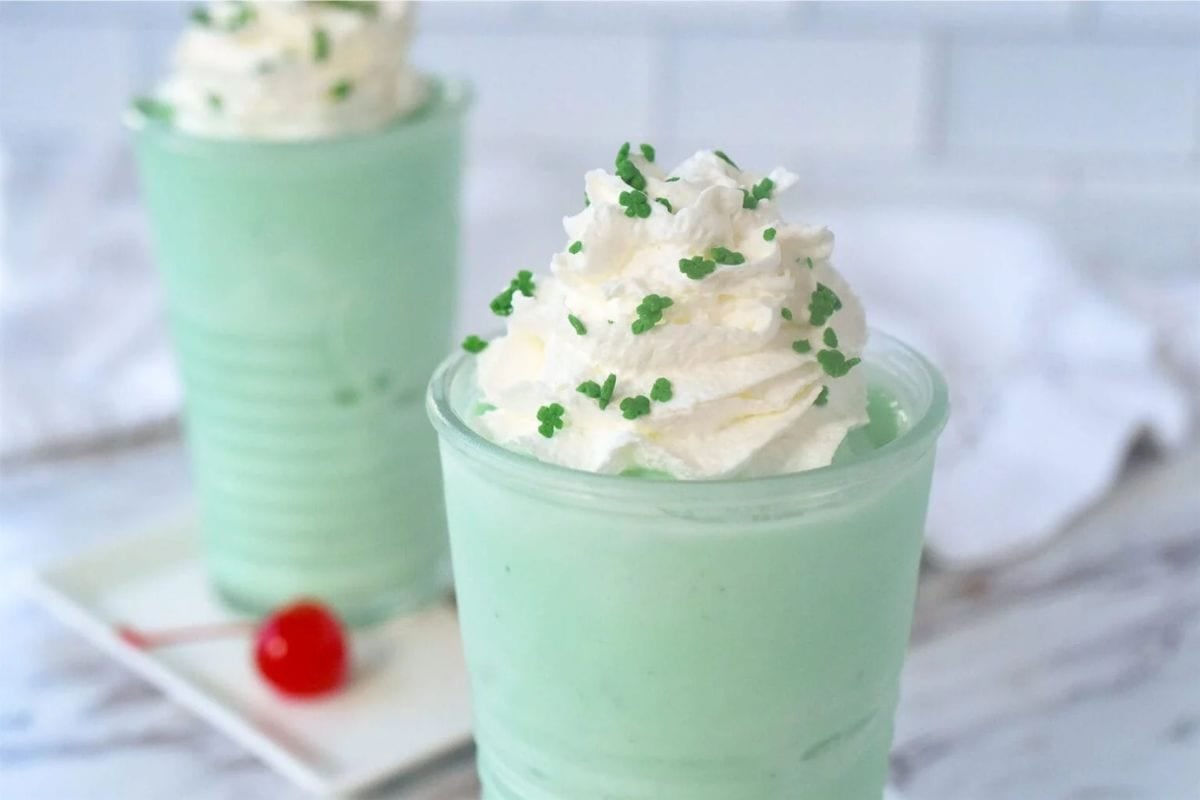 Close-up of a creamy mint dessert topped with whipped cream and green sprinkles in clear cups, with a cherry on a plate in the background.