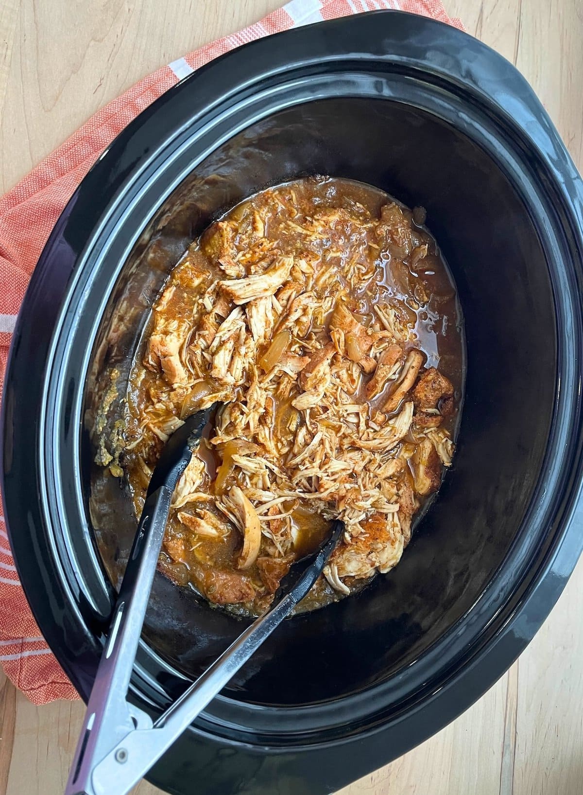 A close-up of shredded salsa verde chicken in a slow cooker with a rich sauce, served with tongs for easy serving.