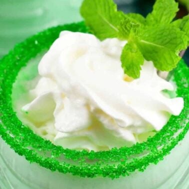 Close-up of a mint green drink topped with whipped cream and garnished with fresh mint leaves, featuring a rim coated in green sugar.