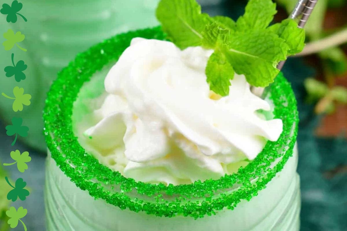 Close-up of a mint green drink topped with whipped cream and garnished with fresh mint leaves, featuring a rim coated in green sugar.