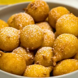 A close-up view of golden-brown fritters dusted with powdered sugar in a white bowl.