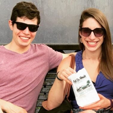 Two smiling individuals enjoy their time together, holding bags of beignets from Café Du Monde while sitting on a bench.