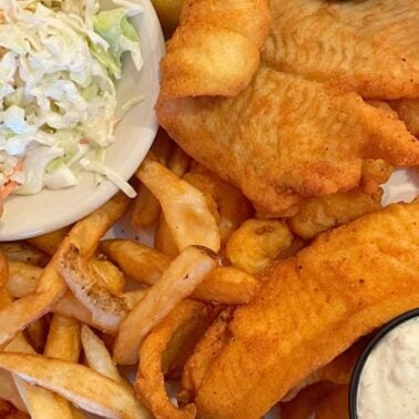 A delicious fried fish platter featuring golden-brown fish fillets, crispy French fries, a side of creamy coleslaw, and a small bowl of tartar sauce.