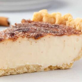 A slice of creamy cinnamon custard pie on a white plate, showcasing a flaky crust and a dusting of cinnamon on top.