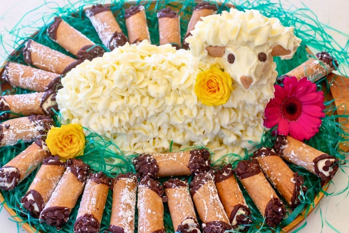 A decorative Easter dessert featuring a lamb-shaped cake made of frosting, surrounded by chocolate-dipped cannoli, and adorned with colorful flowers and green grass.