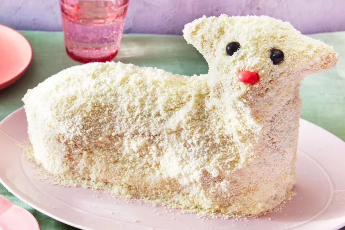 A charming lamb-shaped cake covered in white frosting and shredded coconut, with candy eyes and a red nose, presented on a pink plate with a glass of sparkling water in the background.