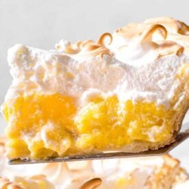 A close-up of a slice of lemon meringue pie being lifted by a serving utensil, showcasing the yellow filling and fluffy meringue topping.