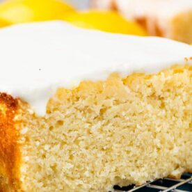 A close-up of a moist lemon cake topped with a white glaze, surrounded by fresh lemons on a cooling rack.