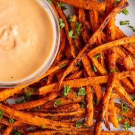 Plate of golden-brown crispy sweet potato fries garnished with fresh herbs, accompanied by a creamy dipping sauce in a bowl.