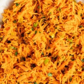 A close-up view of a vibrant shredded carrot salad mixed with green onions and herbs in a white bowl.