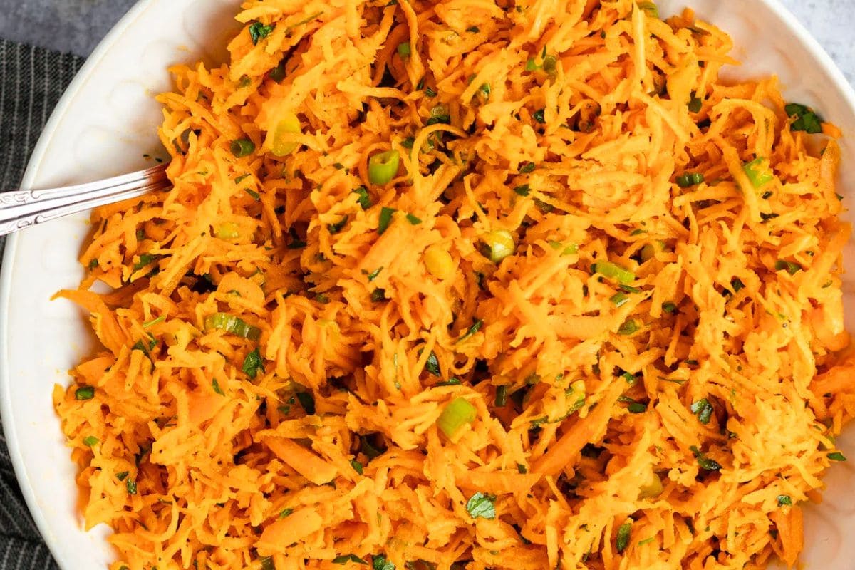 A close-up view of a vibrant shredded carrot salad mixed with green onions and herbs in a white bowl.
