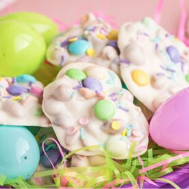 A vibrant basket filled with colorful chocolate candies and plastic Easter eggs surrounded by bright pastel grass.