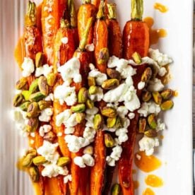 A vibrant dish of roasted carrots topped with crumbled goat cheese and garnished with pistachios, drizzled with a sweet sauce, on a white platter.