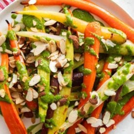 A vibrant plate of roasted rainbow carrots topped with sliced almonds and drizzled with green pesto, presented on a white plate with a red and white striped cloth in the background.