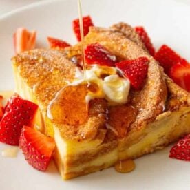 A slice of French toast topped with syrup, whipped cream, and fresh strawberries on a white plate.